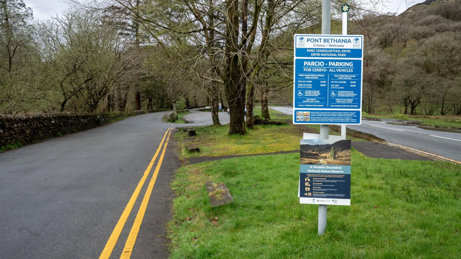 A car park is pictured with road markings, trees and grass by it. A sign listing the parking rules and fees is on a pole.
