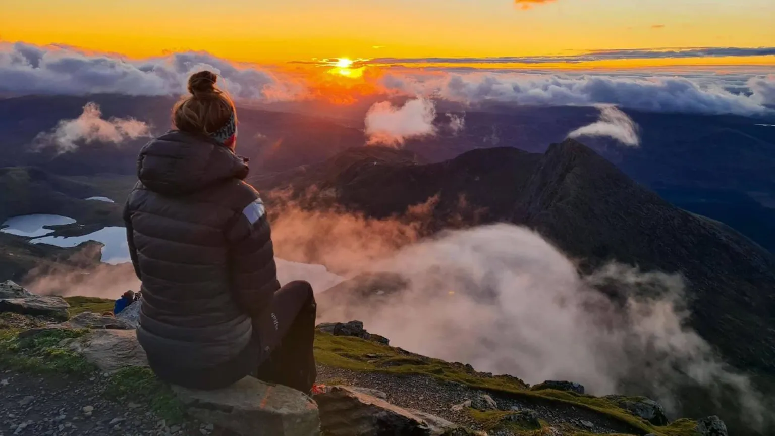 Gemma McInnes A woman is sat on a stone looking at a vast mountainous landscape at sunrise.
