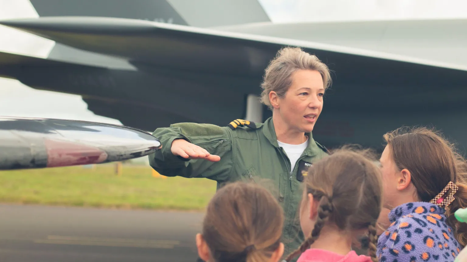 TECgirls The picture shows a group of young people standing on an airfield, gathered closely around an adult who is wearing a green flight suit. The adult has one arm raised and held flat, as if explaining or pointing something out. Behind them is a large military aircraft parked on the tarmac. The aircraft has a dark grey body, angular lines and wide wings, with part of the fuselage and tail section clearly visible.