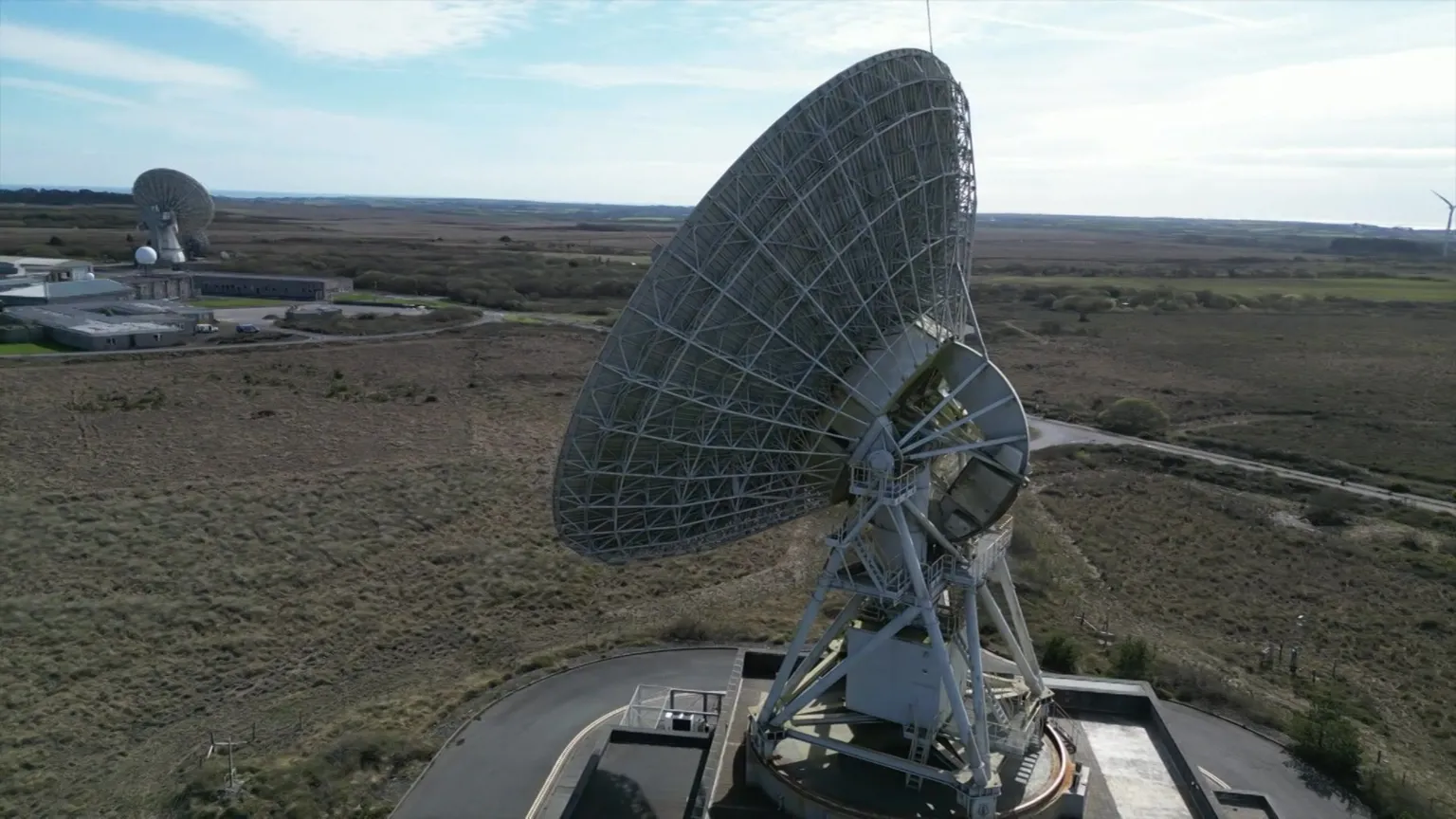 Goonhilly Earth Station The picture shows a very large parabolic satellite dish, also known as a radio or communications antenna, mounted on a heavy steel support structure. The dish surface is made from a metal lattice rather than a solid sheet, and it is angled upwards as if tracking signals in the sky. At the centre of the dish there is a feed horn and receiver assembly, held in place by metal struts that connect back to the main structure.