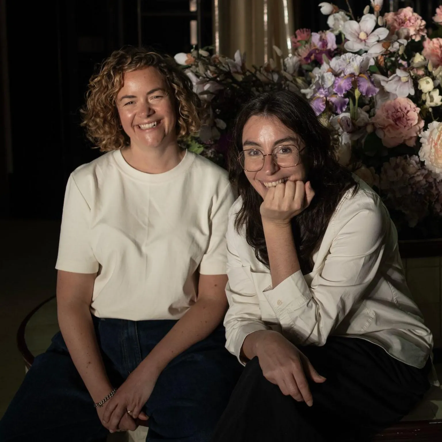 Jon Pountney/WNO Two women wearing white tops and dark trousers smile broadly at the camera. It's a formal pose but they look relaxed. They are sitting in front of a large theatrical bouquet of flowers. Sarah on the left is sitting with her arms on her legs, but Adele on the right is leaning forward and resting her chin on her left arm. 