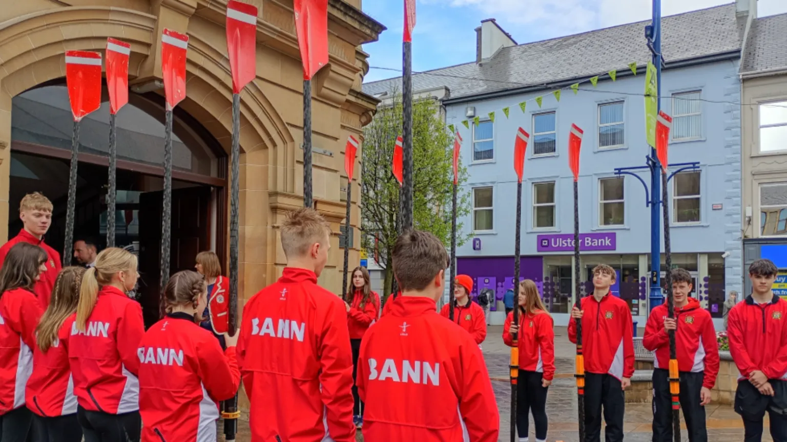 Members of Bann Rowing Club forming a guard of a Guard of Honour outside Coleraine Town Hall. They are wearing red coats and holding padels. 
