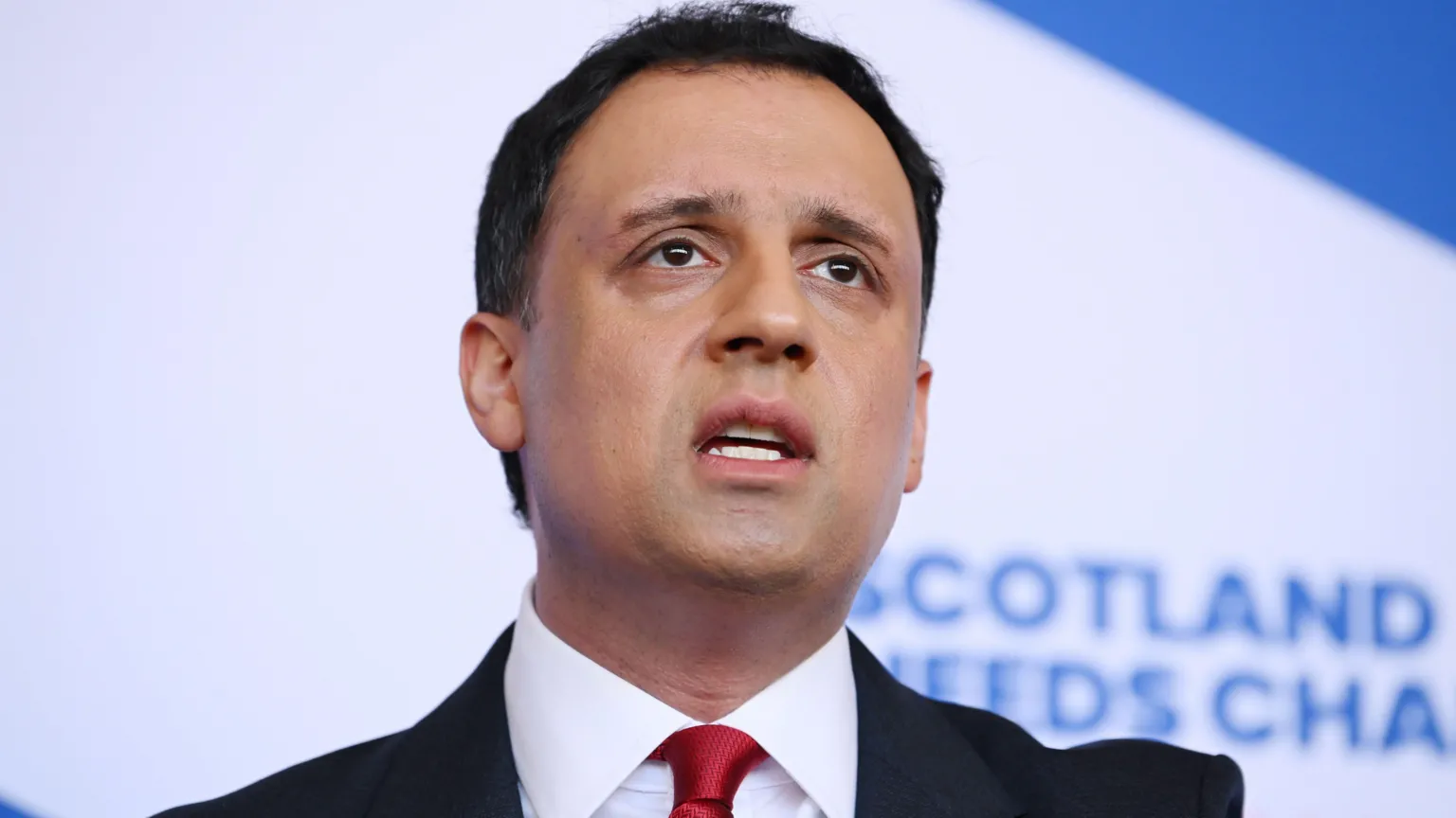  Anas Sarwar is looking sternly at the camera. He has short, dark hair and is wearing a brown jacket and a white shirt. There are campaigners and houses behind him