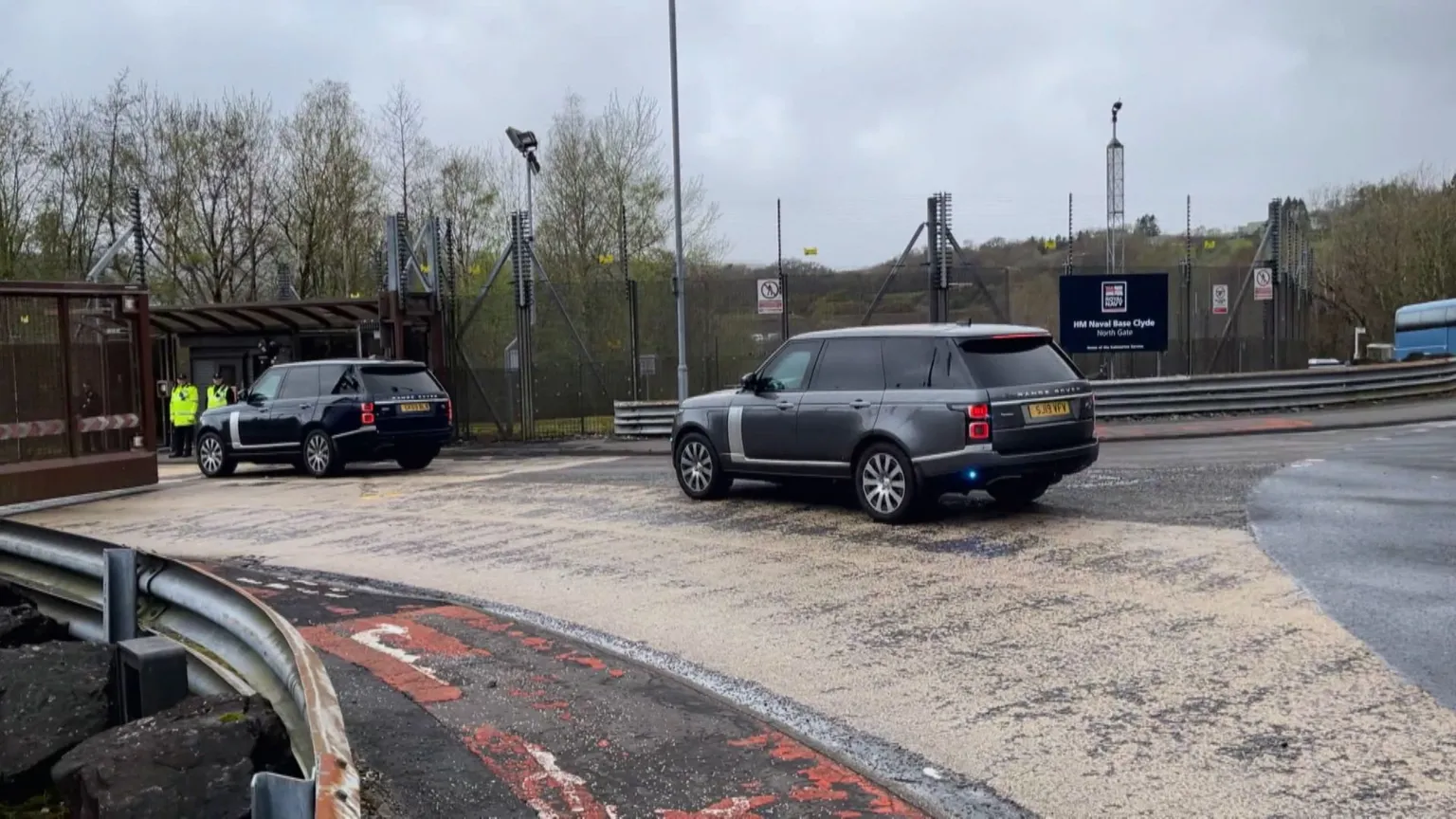 Two black cars driving into the entrance of the Faslane nuclear base. There are police officers or security guards at the entrance wearing hi-vis jackets. 