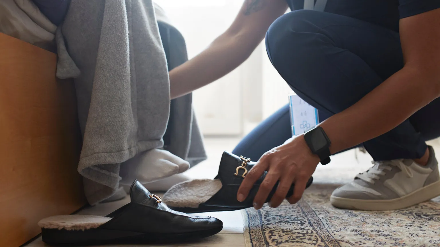  Photo taken from the floor of a care worker helping a person wearing socks and a dressing gown who is sitting down. The worker is holding a black show with a gold buckle. 