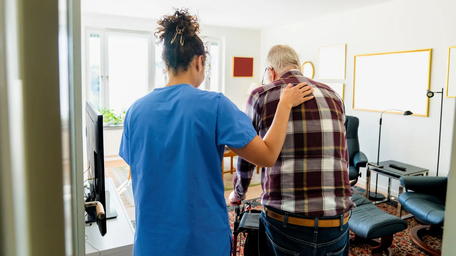  A homecare worker is has a hand resting on an elderly man's back. He is using a zimmer frame to walk. The worker is wearing blue overalls and the man is wearing a maroon and yellow checked shirt and jeans. They are in a living room. There's a TV, some tables and a chair in the background. 