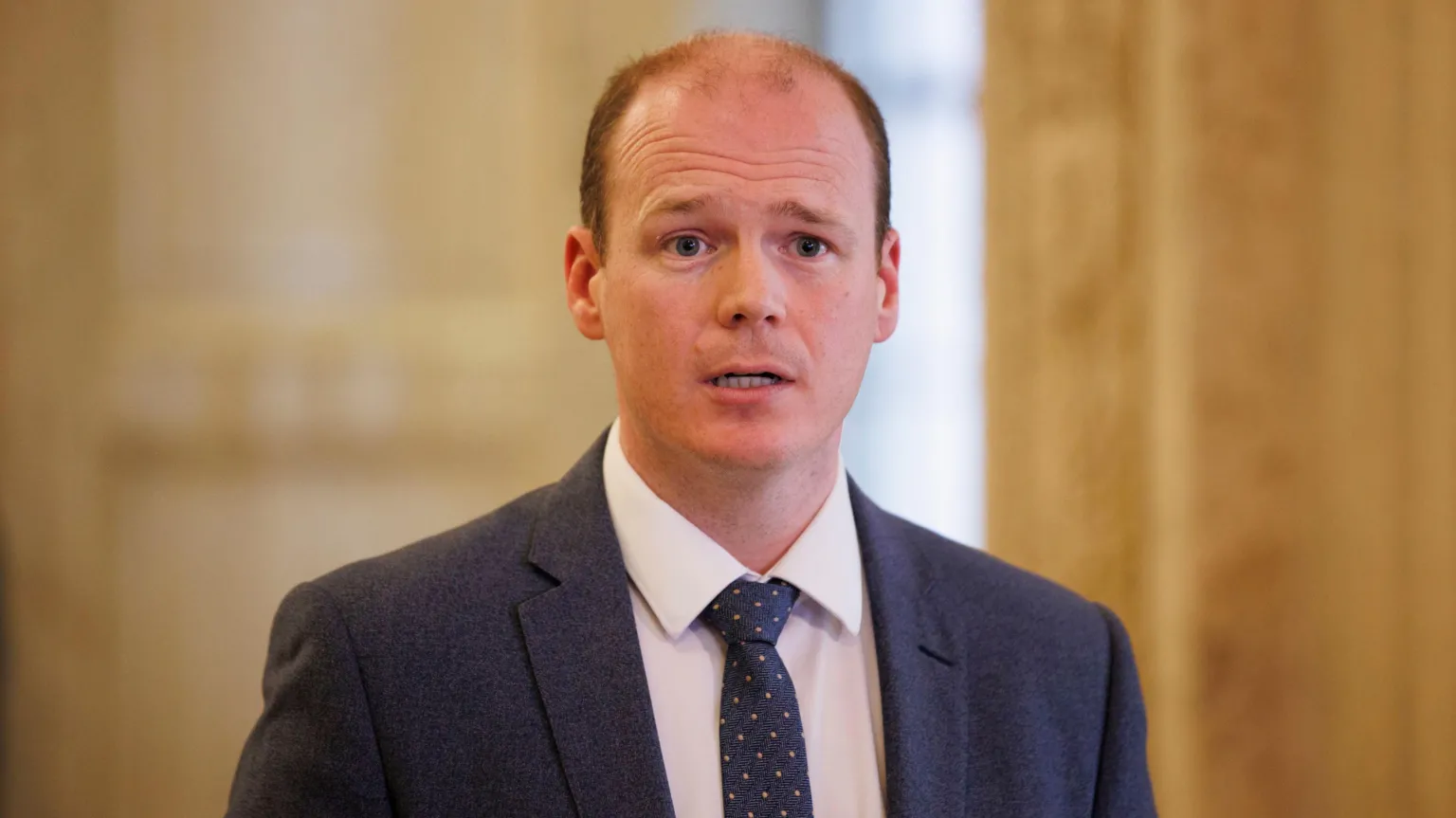  Lyons is wearing a navy suit with a white shirt and a navy tie with pink spots. He's talking. The background is out of focus. 