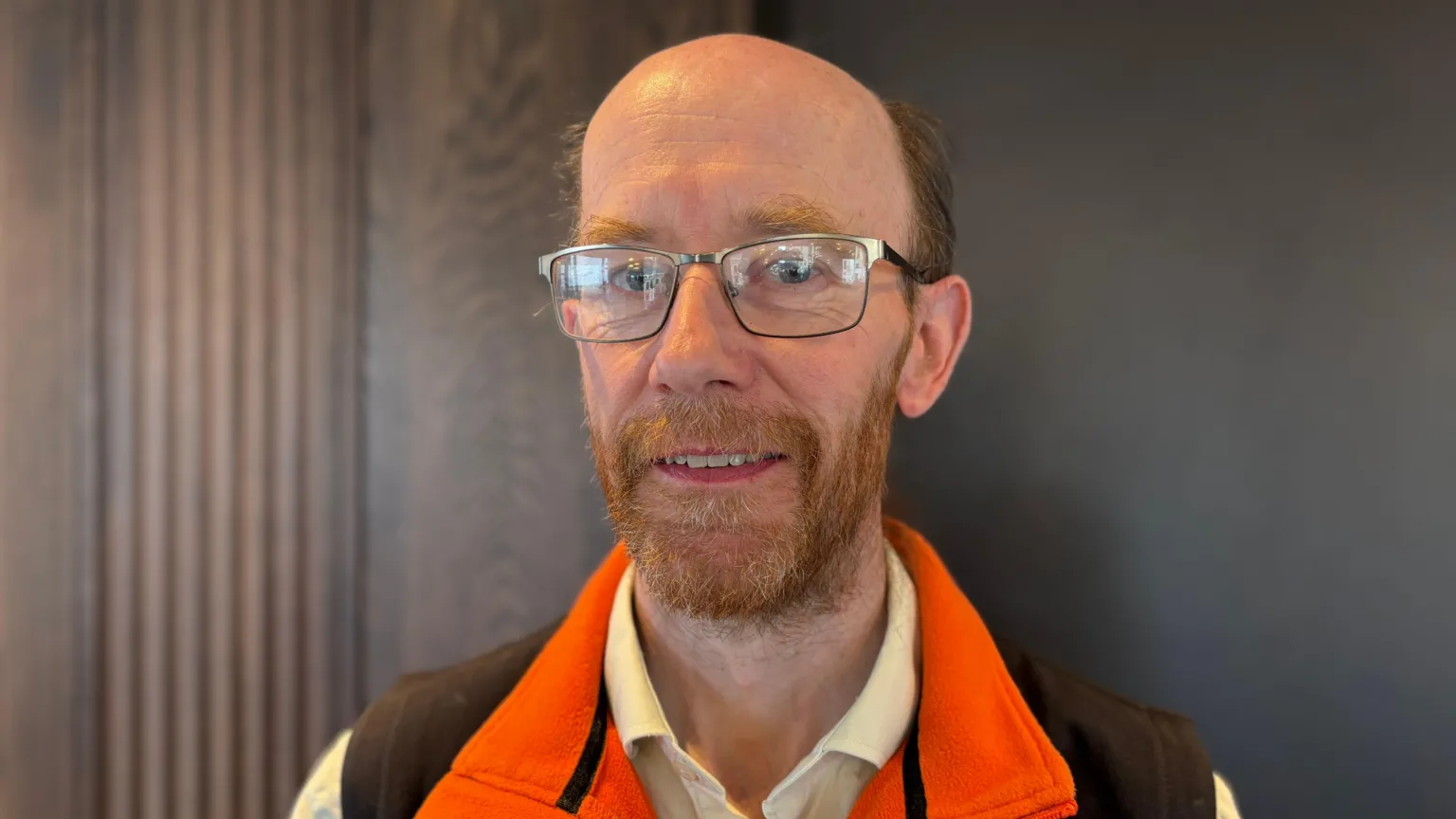 A man stands in front of a wooden wall. He is bald, with glasses and has a red beard. He's dressed in a pale yellow polo shirt with an orange and brown body warmer over it. He is smiling. 