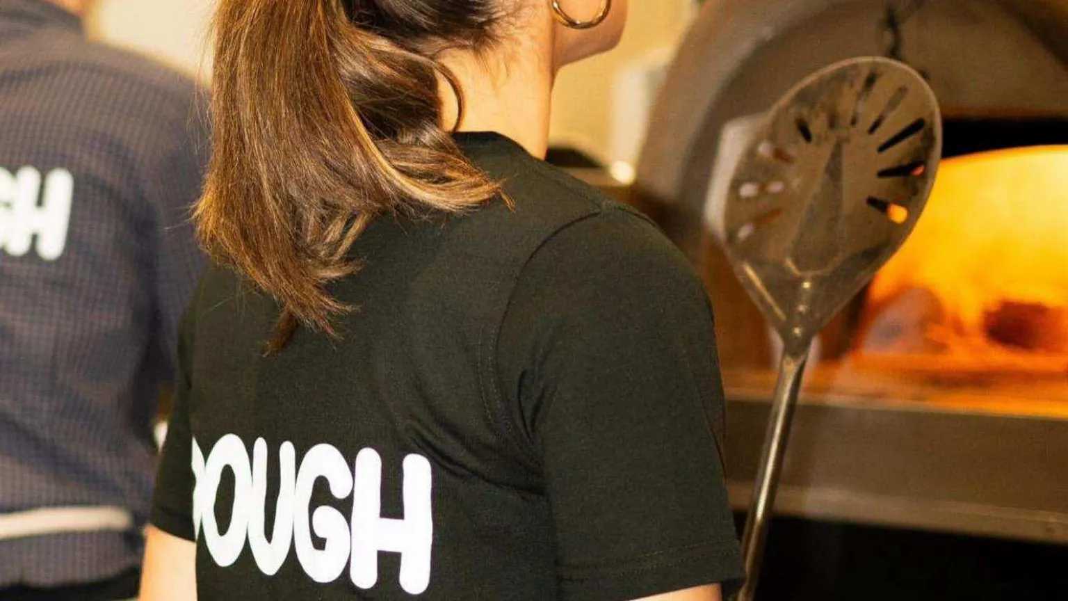 Jamie Watkins A young female wearing her hair in a ponytail and in a black T-shirt with 'DOUGH' written in white capital letters on the back. She's stood facing a pizza oven in the store.