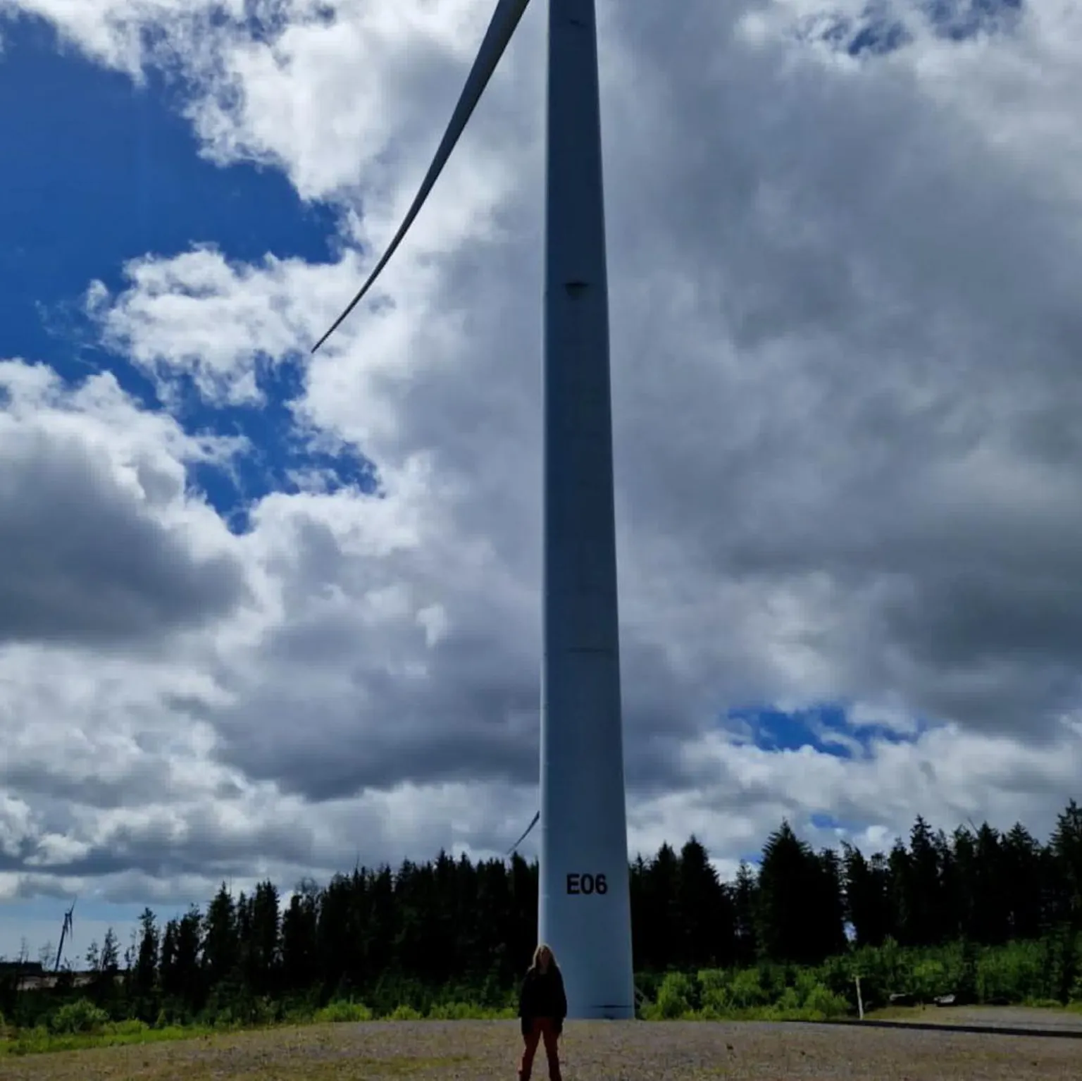 Grace Lloyd A person standing at the base of a tall wind turbine near Treorchy set against a cloudy sky, with trees and open ground in the background. Grace appears small in scale compared with the turbine, highlighting its height and the surrounding landscape.