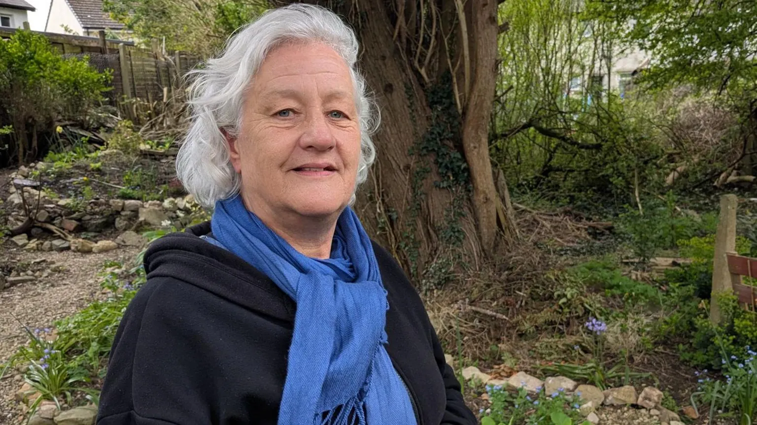 Cathy is standing in a garden area with trees, shrubs and stone borders, in west Cardiff, wearing a dark top and a blue scarf. Spring plants and greenery surround the scene, with houses and garden fencing visible in the background.