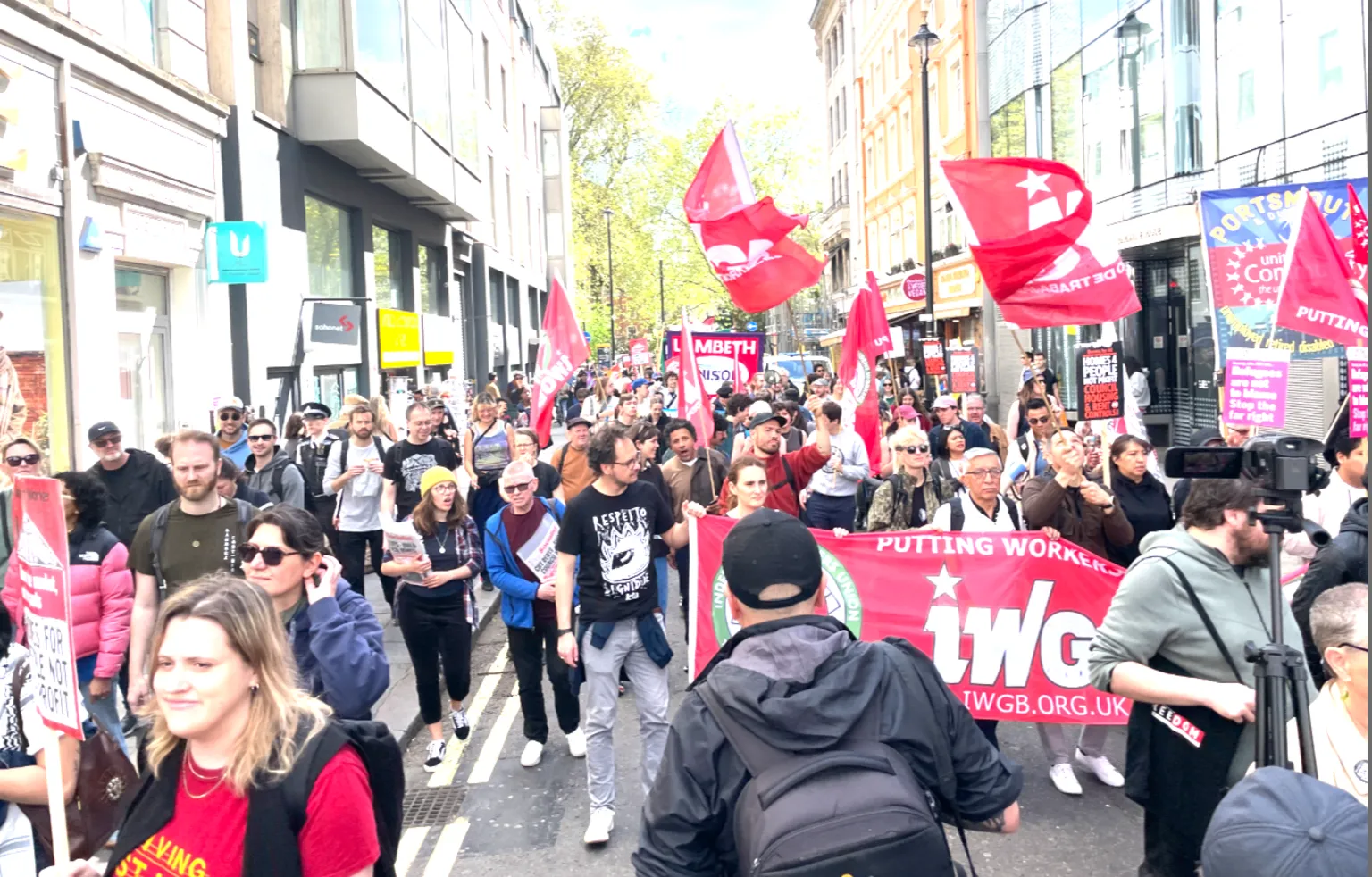 Demonstrators walk down a city street carrying red flags and a banner, with shops and buildings lining the road