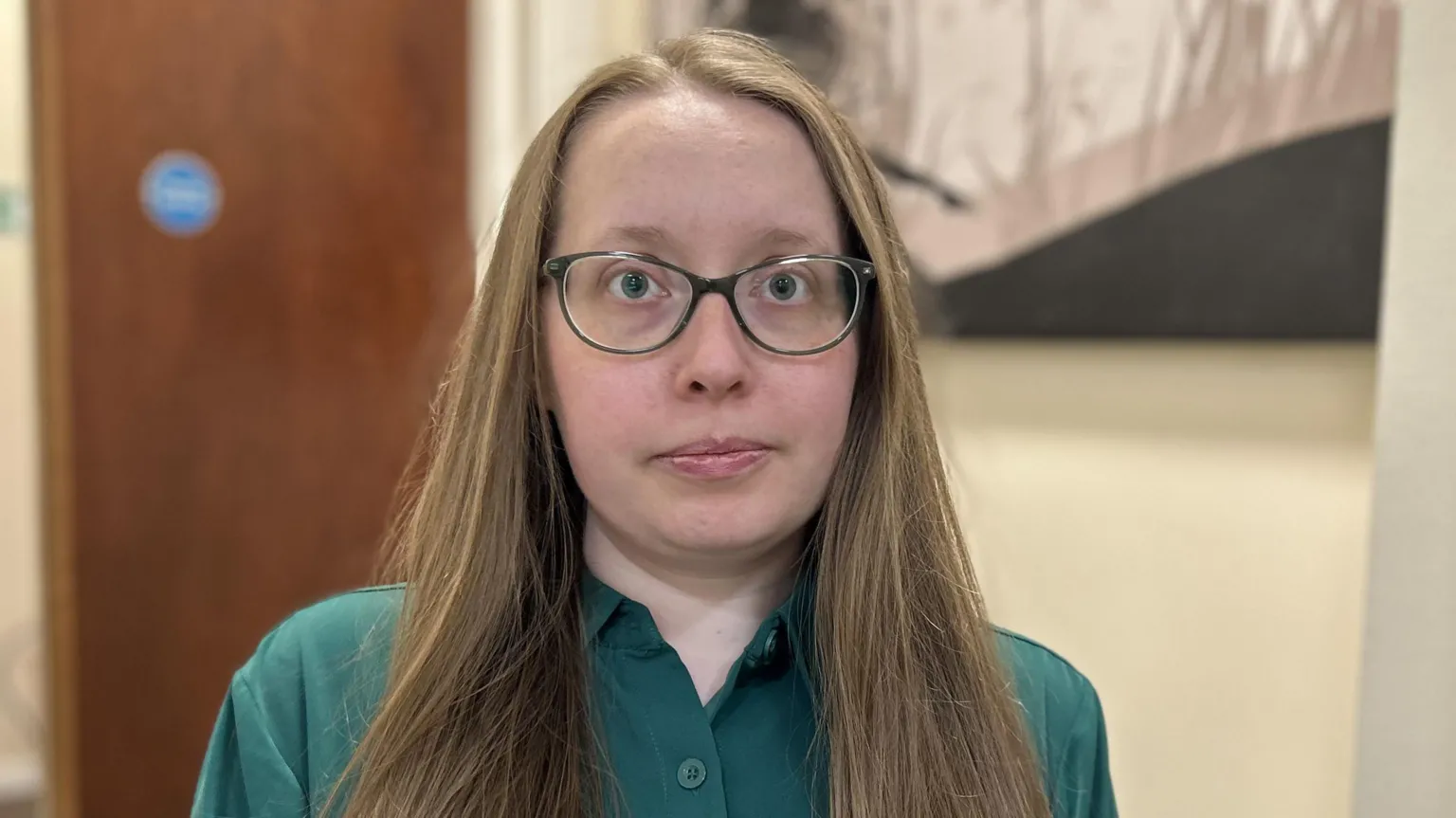 A woman with long dark-blonde hair who is wearing dark-rimmed glasses looks at the camera. Her hair falls to the front over her dark green blouse which is unbuttoned at the neck