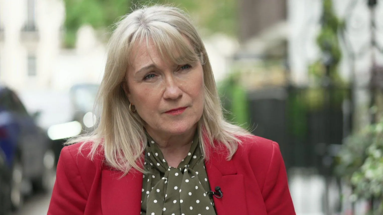 A middle-aged woman with shoulder-length hair looks out of the frame to the right (her left). Behind her, out of focus, are railings, some greenery and a parked car. She is wearing a bright red jacket and a green-and-white spotted scarf
