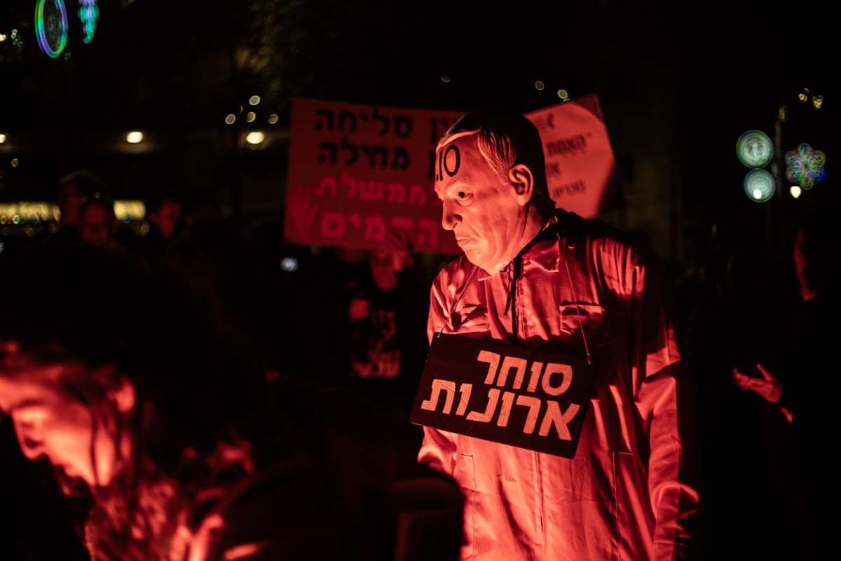 Protesters, including one with a Netanyahu mask, are lit red at night. The protester with the Netanyahu mask holds a sign in Hebrew. 