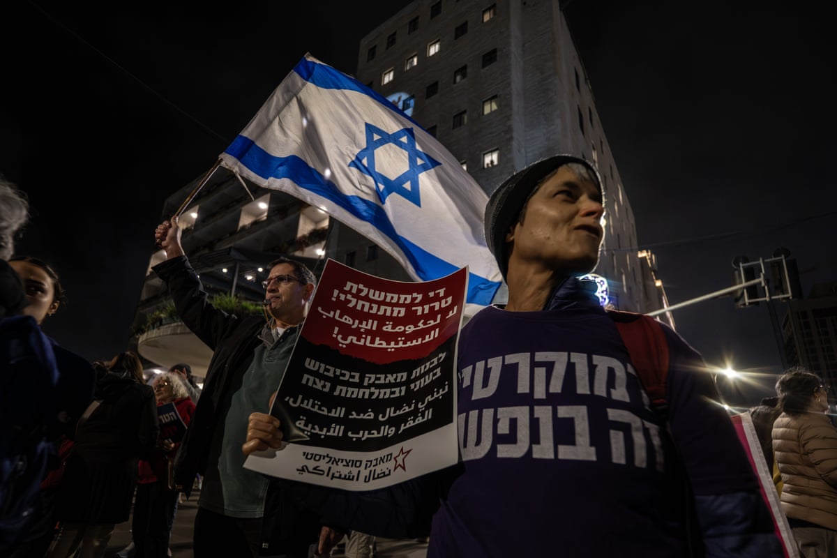 A protester holds a sign in Hebrew while to her right, a man waves an Israeli flag. 