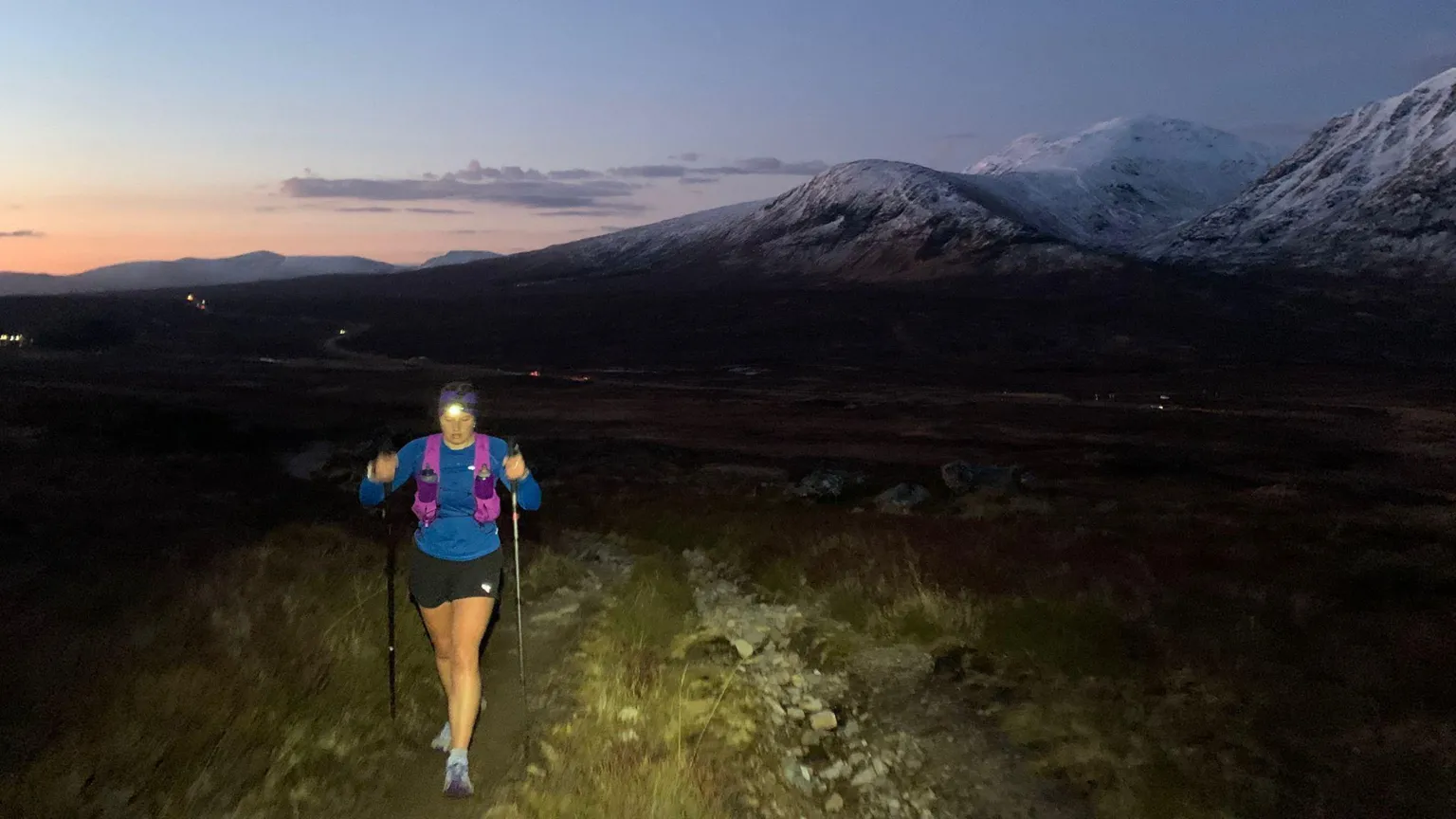 Freya Shepherd Freya running at night, she is wearing a blue, long-sleeve top, black shorts , magenta backpack and a flash light on her forehead. She is holding walking sticks in her hands. Mountains covered in snow are in the background. It is dusk and the sunset is visible in the background.