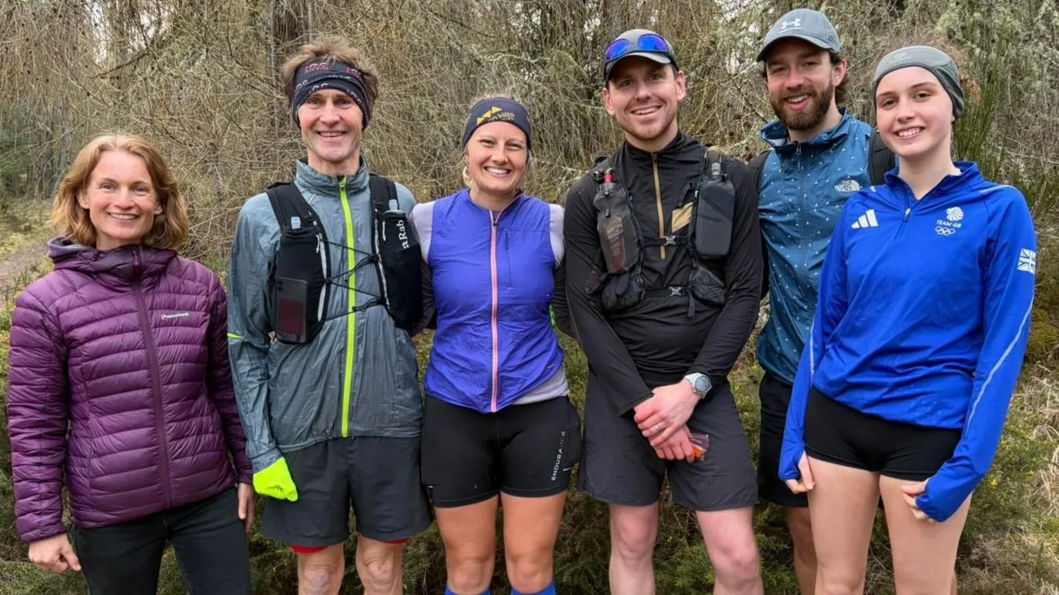Freya Shepherd The photo taken in the woods, the trees are visible on the background. Freya is standing in the middle of the shot and her support runners are standing next to her. On her right hand side is a man in a grey running jacket and shorts, with black headband, and a woman with shoulder length fairly red hair, purple jacket and black leggings. On Freya's left hand side is a man in black top and shorts with a grey cap, next to him on his left stands another man in a blue jacket, grey cap, black shorts and his having a beard. And then, further left stands a woman in blue adidas quarter zip top, black shorts and grey headband. Everyone on the picture has big smile on their face.