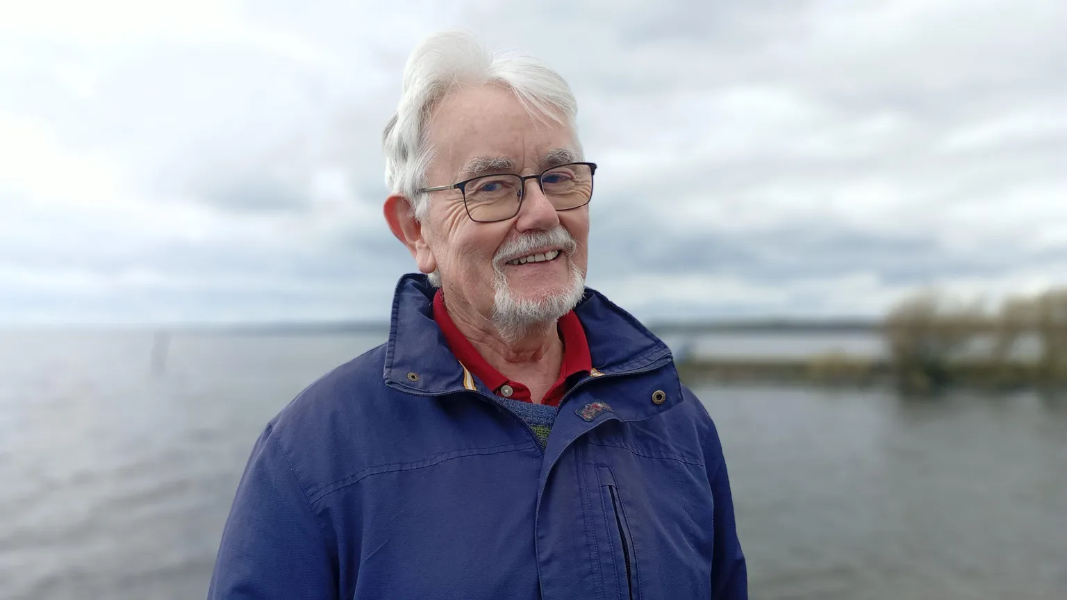 Paddy smiling at the camera. He has grey hair and is wearing glasses and a red polo shirt with a navy jacket zipped up over it.