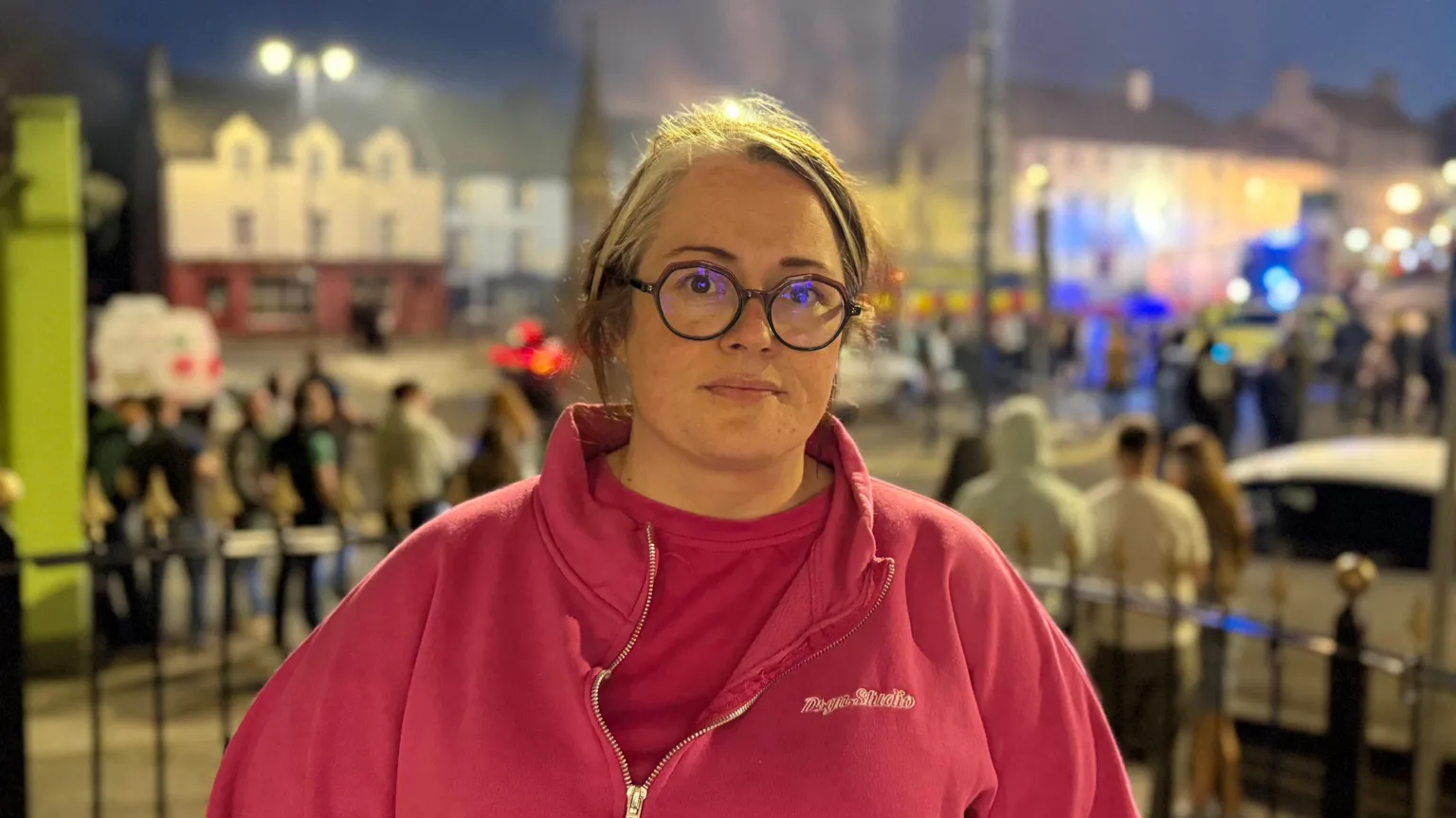 Sian Mulholland standing in front of the scene looking at the camera. She is wearing a pink top and fleece and round glasses. Her hair is tied back. In the background, there are small crowds of people standing in the street.