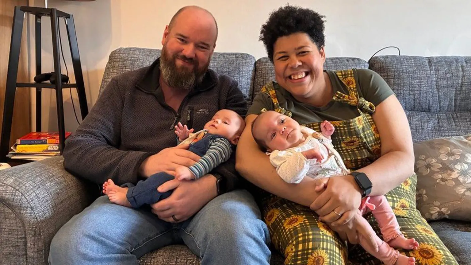 Elita and Adam sit on a couch, each holding a baby. Adam wears a dark jumper, and Elita wears a bright sunflower-patterned overalls. A lamp and stacked books rest on a small table beside them, creating a warm, homey scene.