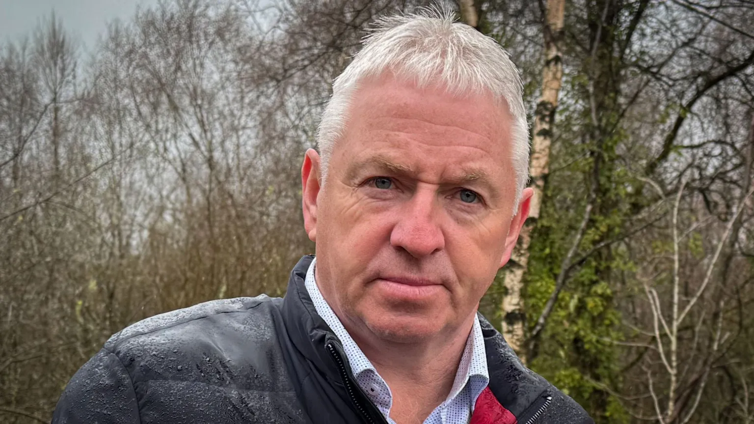 A man in a rain-speckled waterproof jacket, with a fluffy microphone attached to the lapel. He is outside and there are birch trees behind him. He has white hair and blue eyes
