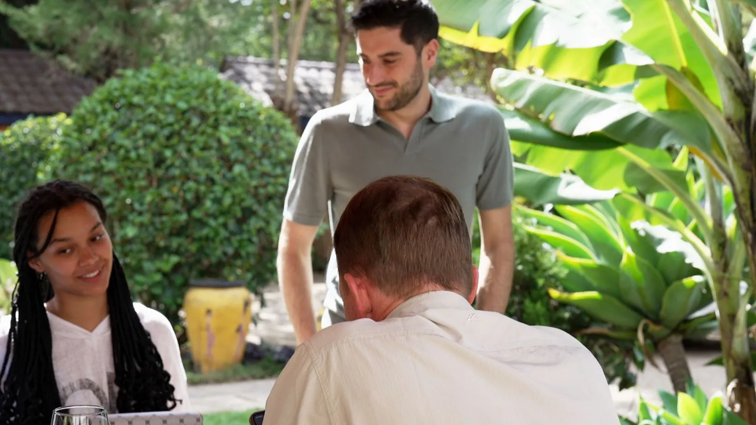 Three people, two white men and a mixed race girl gather around a table with green plants and bushes in the background 