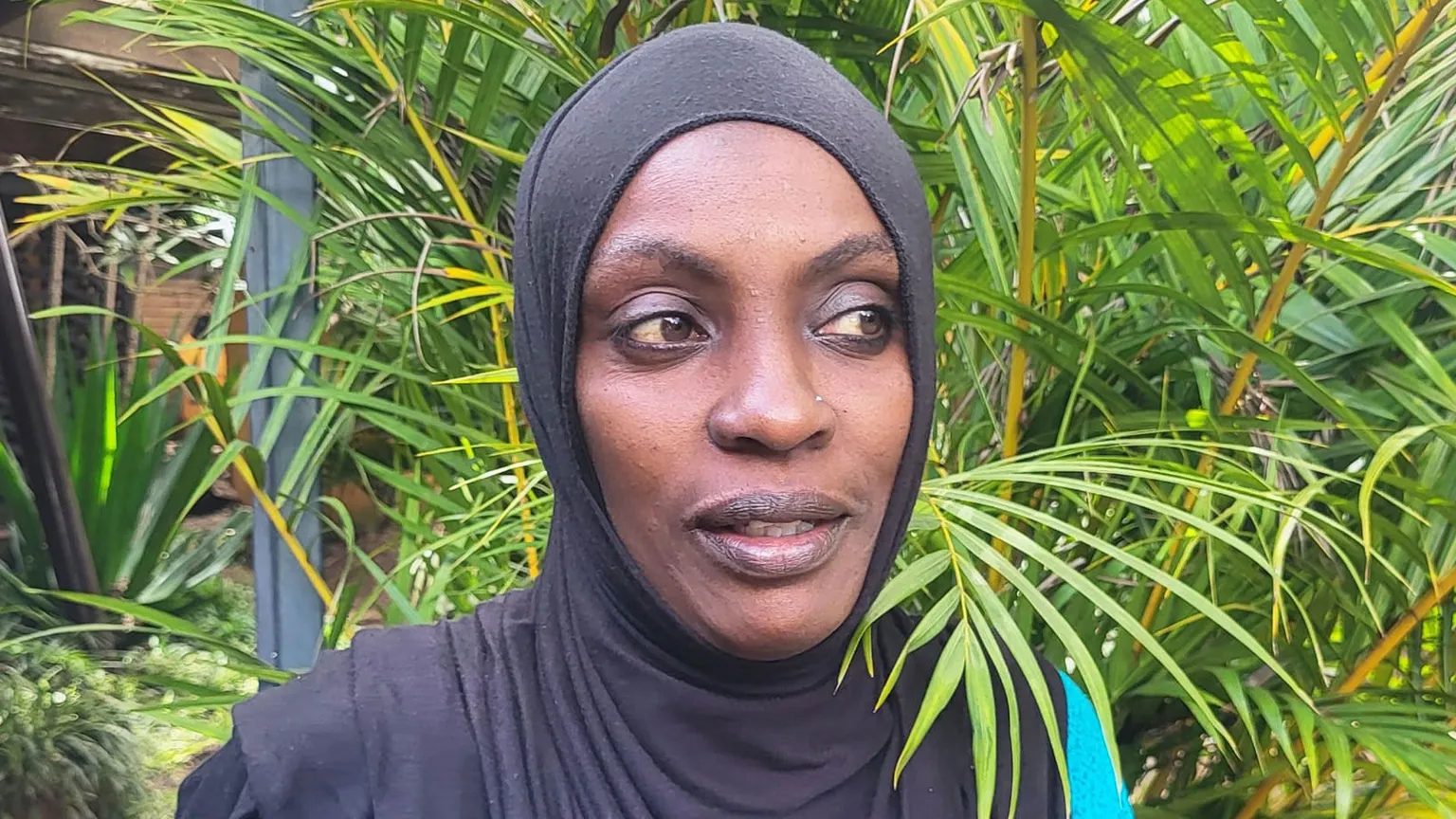 Nasibo wears a black hijab and is photographed with green plants behind her - she is looking away from the camera 
