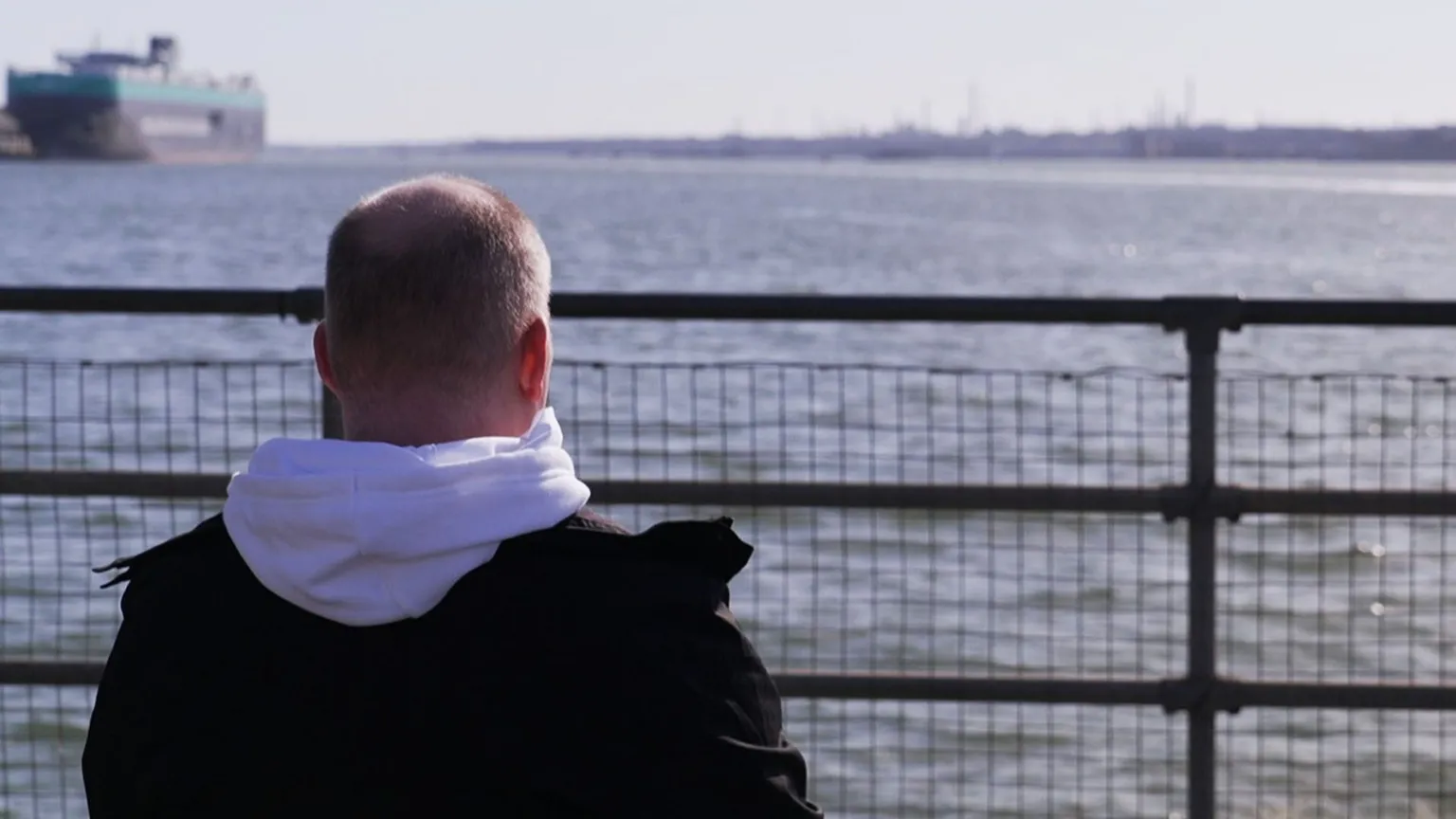 Phill is pictured from behind, sitting and looking out to sea, with a boat in the background. He has short fair hear and is wearing a white hoodie and a black coat.