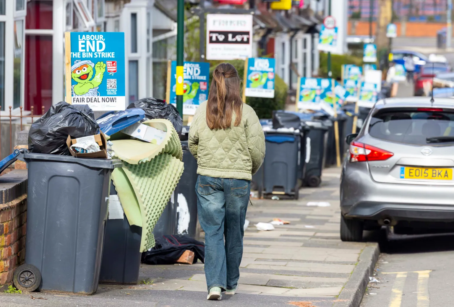 Mark Thomas A woman with long brown hair is walking down a residential street which is lined with overflowing wheelie bins. She is wearing blue jeans and a green padded jacket. Also lining the street are signs someone has put in the front yard of terraced houses, which are blue and say 'Labour, end the bin strike - fair deal now' in white writing.