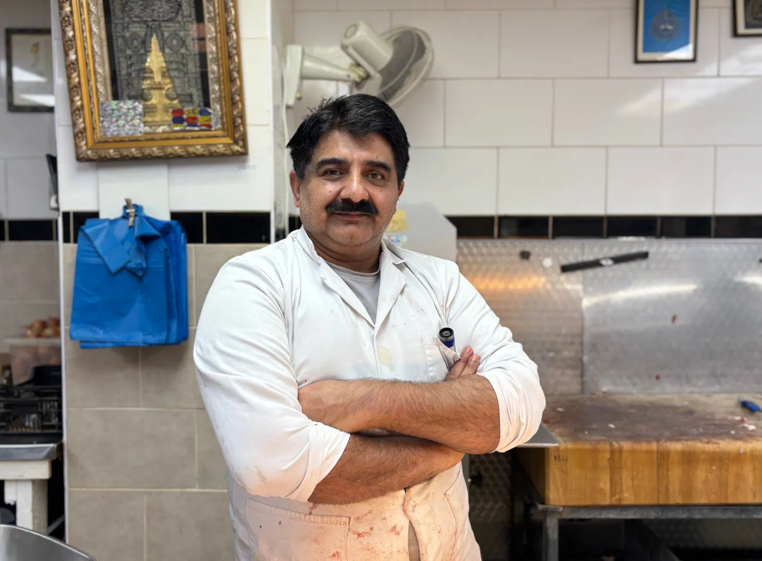 A man with black hair and a thick black moustache is behind the counter of a butcher's shop, he is wearing a white tabard and has him arms crossed and is looking directly at the camera. Behind him is the counter for chopping meat with blue carrier bags on the wall.