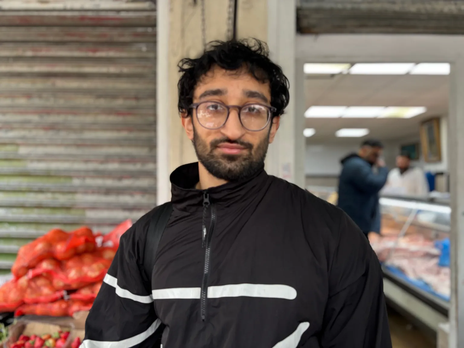 A man with black, short wavy hair, round glasses and a short beard and moustache looks directly into the camera. He is wearing a black zip up coat which has two white stripes across the middle. Behind him is the blurred image of a meat shop