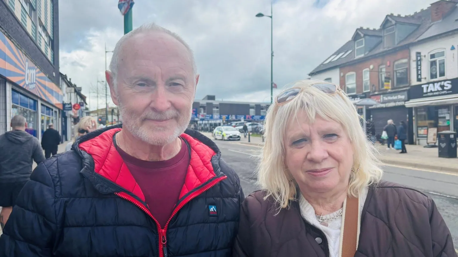 A man and woman stand together looking at the camera. He has short grey hair and a moustache, and wears a black padded coat with a red lining and a maroom jumper underneath. His wife has blonde shoulder length hair and sunglasses on her head. She has a brown padded coat on, a cream jumper underneath and a necklace. Behind them is a row of shops and a main road.