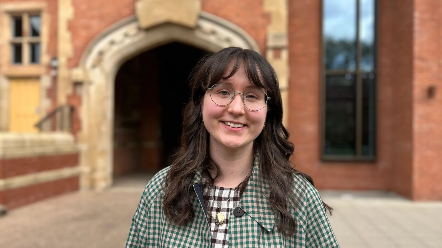 A picture of a woman with dark hair and glasses smiling while wearing a green checked coat. She is standing outside a red brick building during the day. 