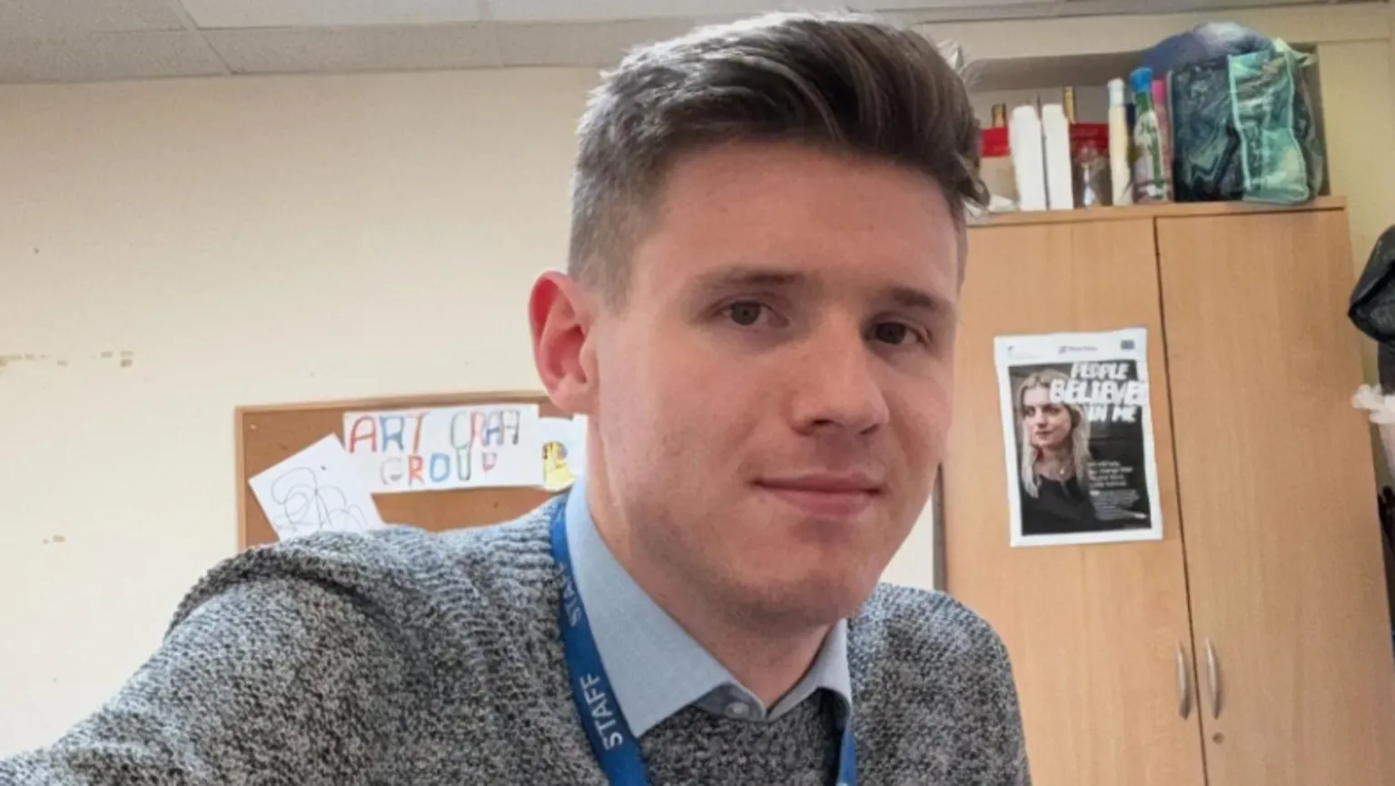 A man wearing a grey jumper and shirt looks towards the camera and smiles while sat inside an office