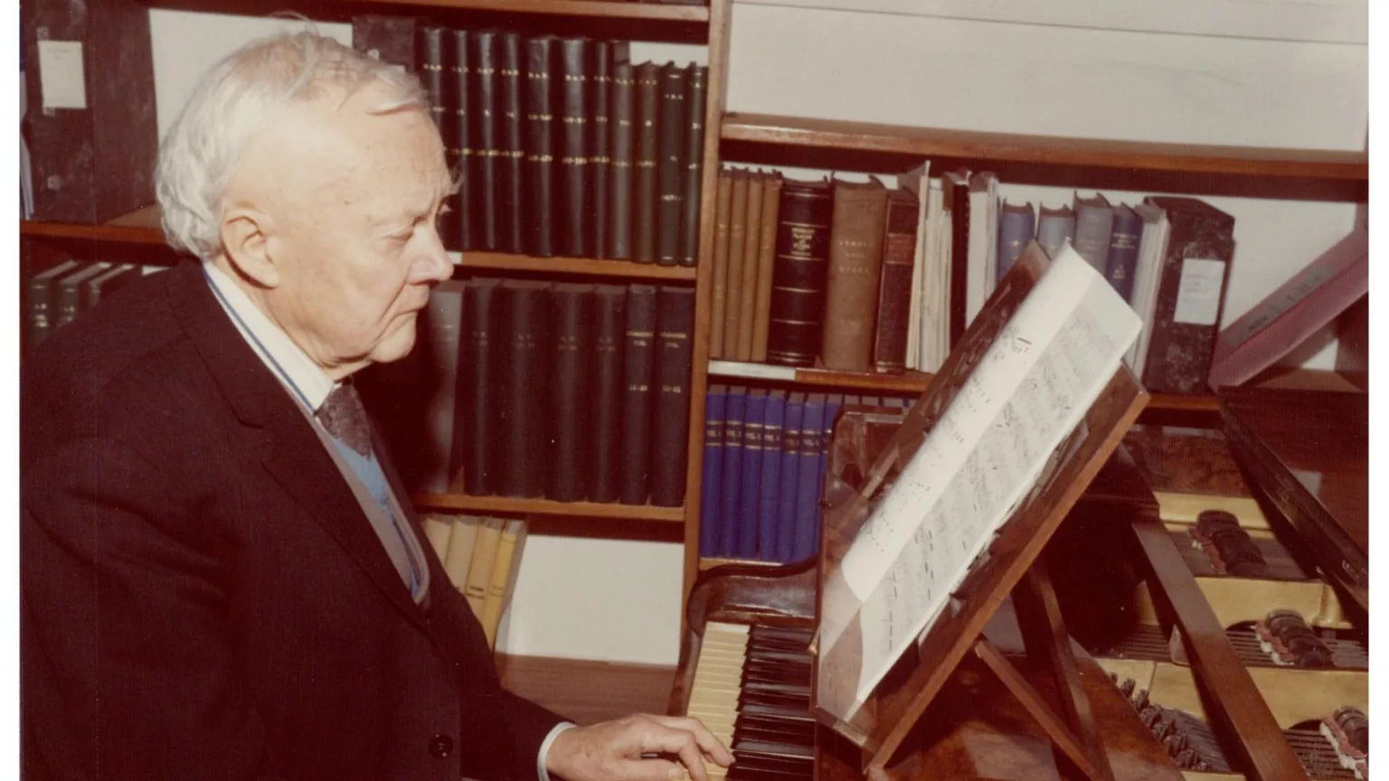 Armagh Planetarium A faded picture of an older man in a suit sitting playing a piano in front of a bookshelf.