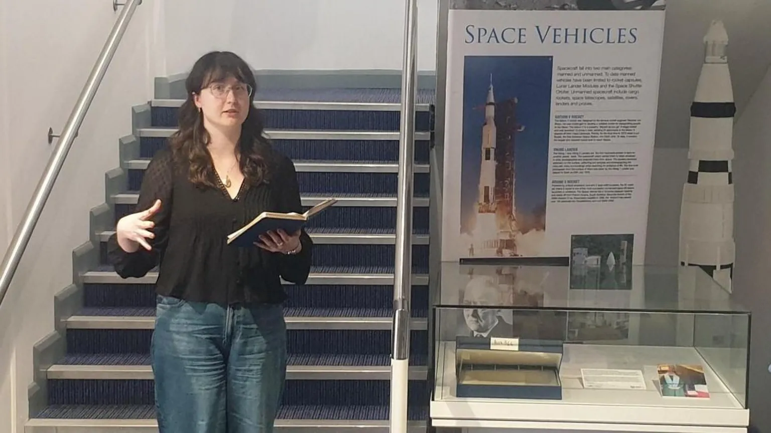 Armagh Planetarium A woman standing speaking with a notebook open in one hand while the other gestures. She has dark hair and is wearing glasses, a black top and jeans. She is standing in front of stairs beside a museum display.