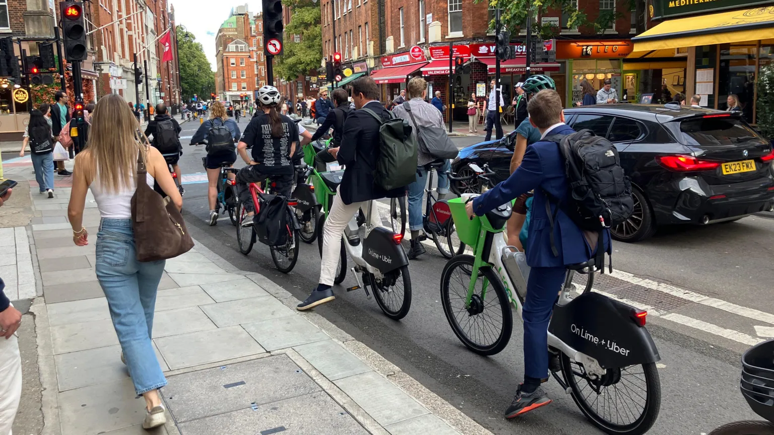 BBC/Harry Low Cyclists, including Lime Bike users, wait at a red traffic light as pedestrians walk past