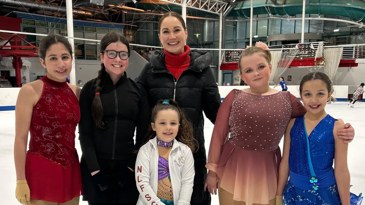 Rhiannon Scott A group of several skaters standing on a rink. They are all children or teenagers, except for Rhiannon Scott, a woman with dark hair and wearing a black jacket with red top. She is standing in the centre of the group. 