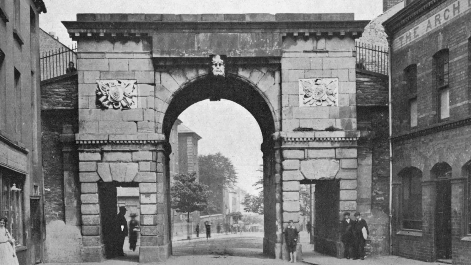 Print Collector//Hulton Archive A black and white archive print of a large stone gate in Derry city centre which forms part of the city's historic 17th century walls. It has a large, arched entrance and smaller rectangular pedestrian entrances on either side of the arch. Several people are standing at the gates including a young, barefoot boy who is leaning against the stone. 