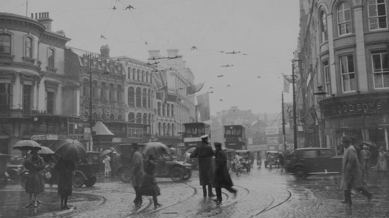 Fox Photos//Hulton Archive A black and white archive photo of a wet and windy day in Castle Place in Belfast city centre, circa 1925. There are tall, ornate buildings lining the cobbled street. Several pedestrians are walking in the rain, some are holding umbrellas. Motor cars and double decker trams are travelling along the street. There is a large union flag flying from one of the buildings.