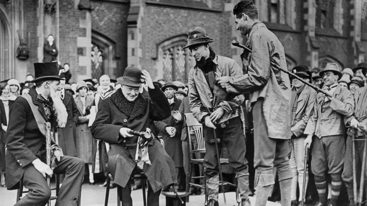 Topical Press Agency/ A black and white archive photo of Winston Churchill visiting Queen's' University, Belfast in September 1926. He is seated on a small stage surrounded by a crowd of onlookers. He is wearing a dark overcoat with a large fur collar. Churchill is being presented with a hat and a clay pipe by students during rag week. One of the students is wearing a top hat and a long, fake beard. 
