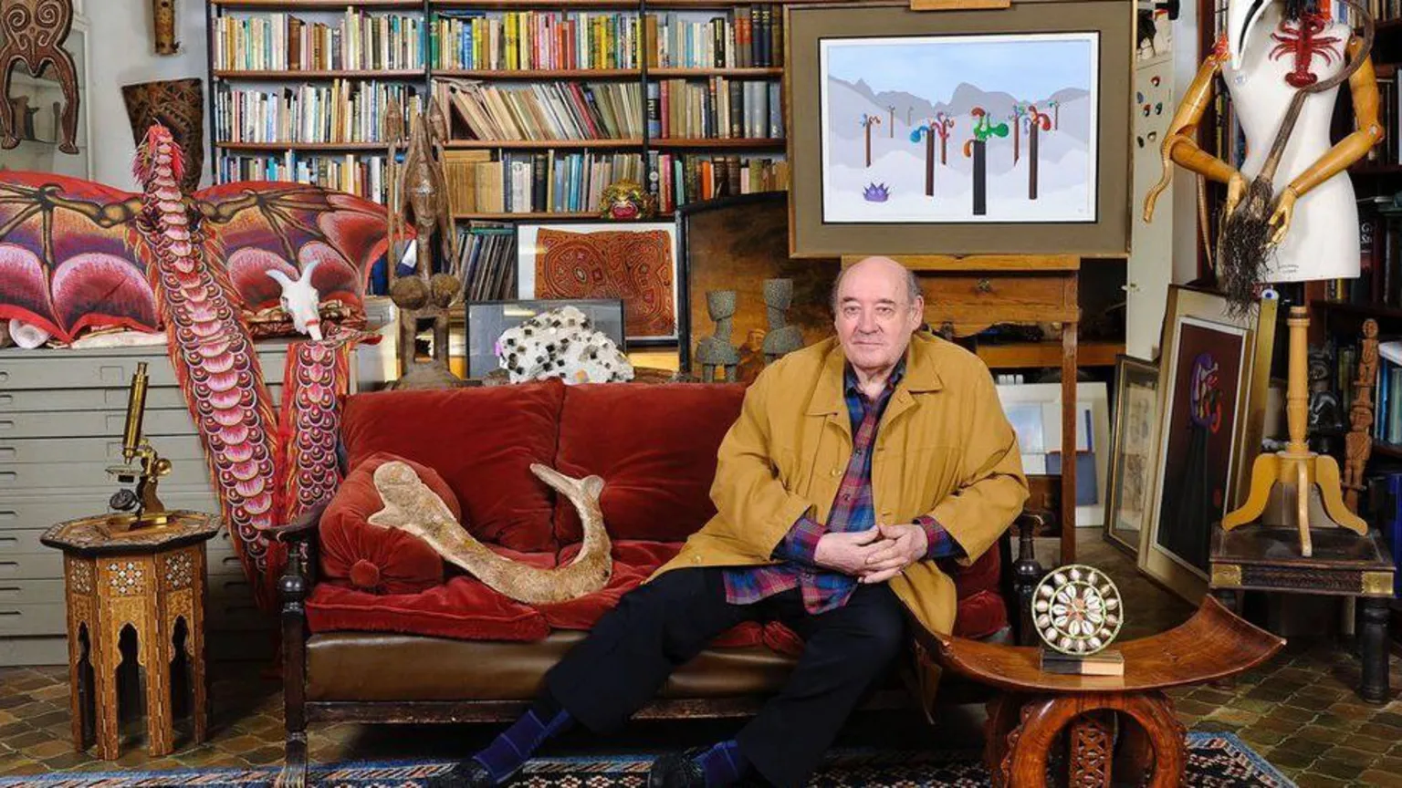 Desmond Morris at home. This looks like a busy sitting room with Morris sitting on a red couch surrounded by books and his art works.