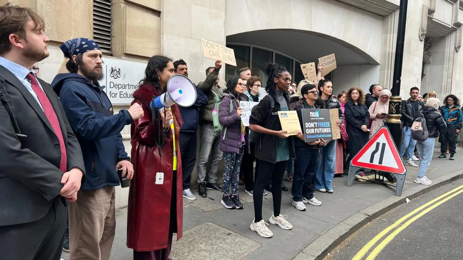 Branwen Jeffreys / BBC A line of protesters stand outside the Department for Education building, some holding placards and one using a megaphone. Visible signs include “Don’t hike our future student loans”. The group lines the pavement beside a road with temporary traffic signs.