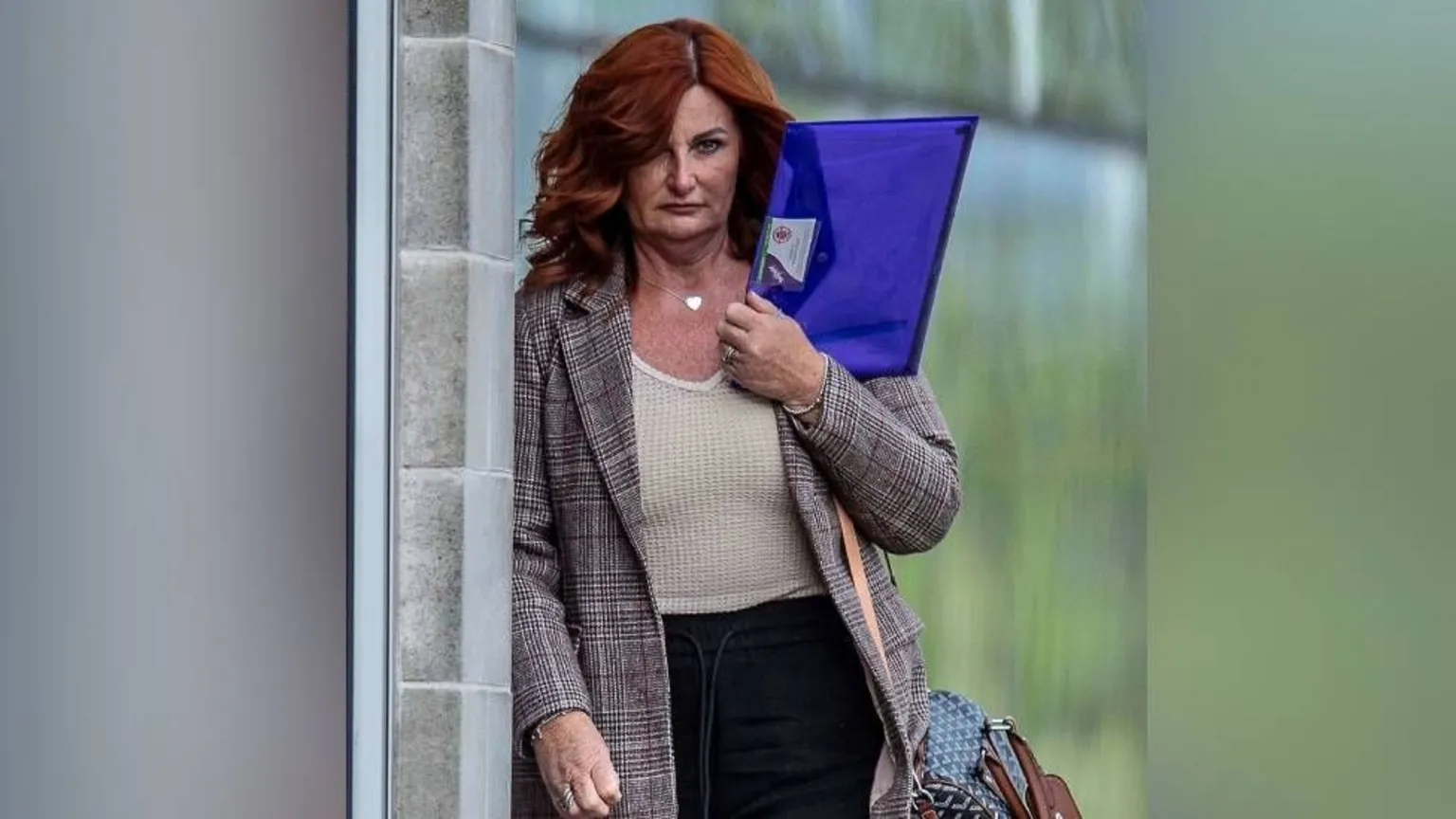 Andrew Price Kershaw is holding a blue plastic wallet with paper inside. She has red hair and is wearing a jacket as she walks by a court.