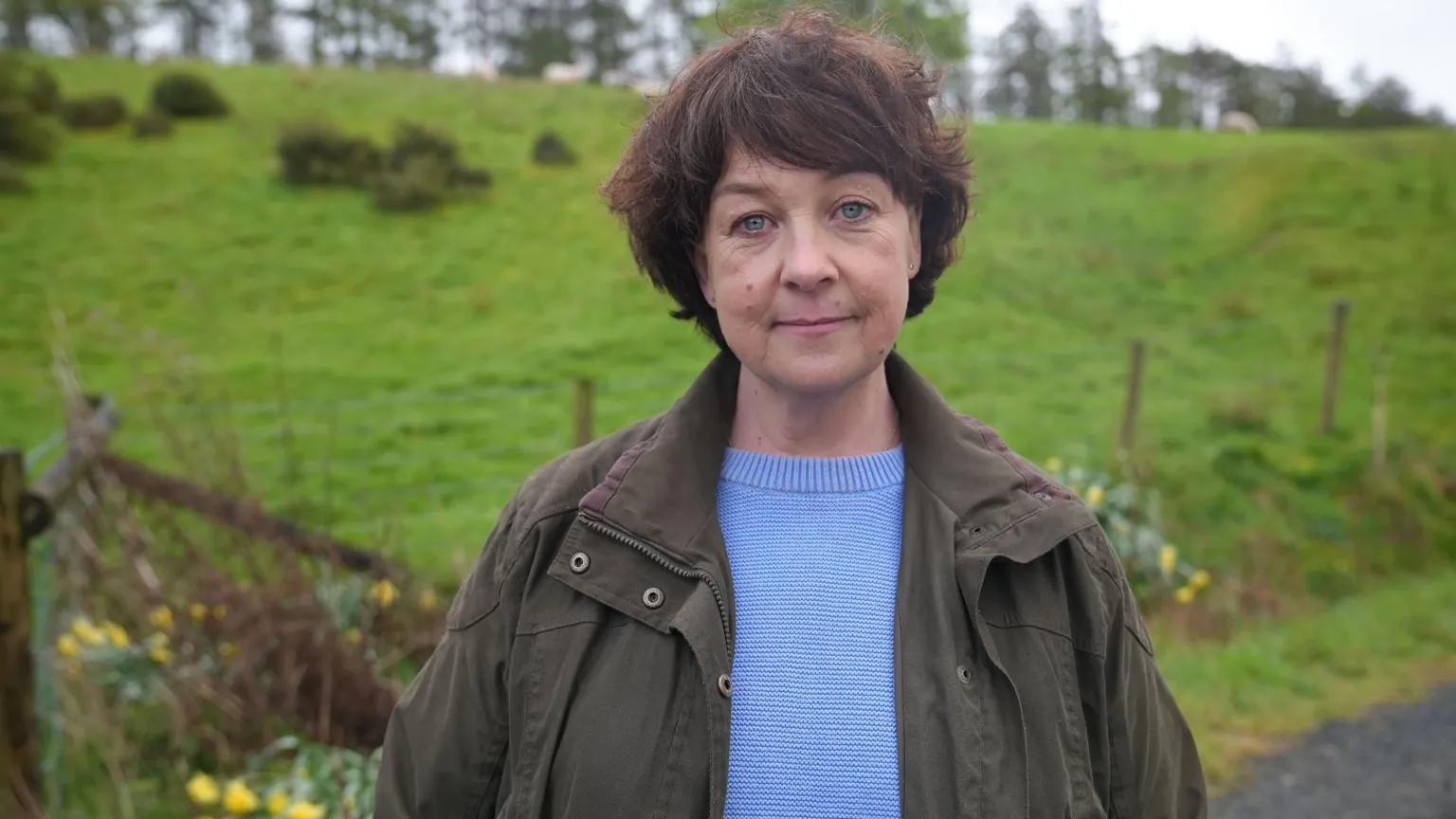 Natalie Barstow, lead claimant, stands on her farm near Builth Wells in Powys. She has brown hair, blue eyes and is wearing a light blue jumper and dark green coat. Green fields and daffodils can be seen behind her.