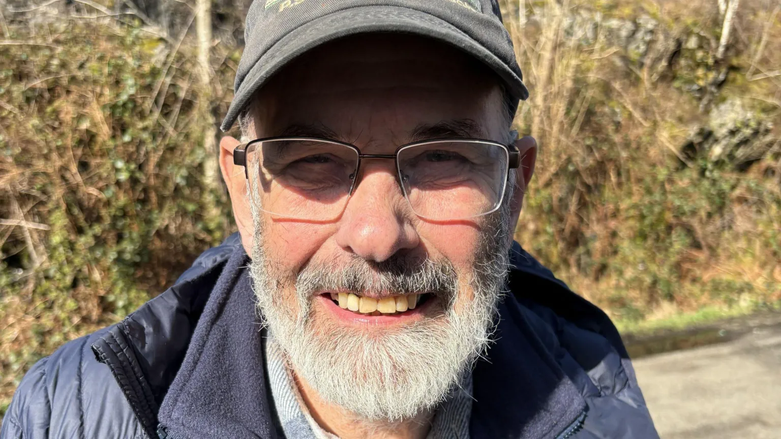 John Urquhart, an older gentleman, with a grey beard and glasses, he wears a navy jacket and a black cap.