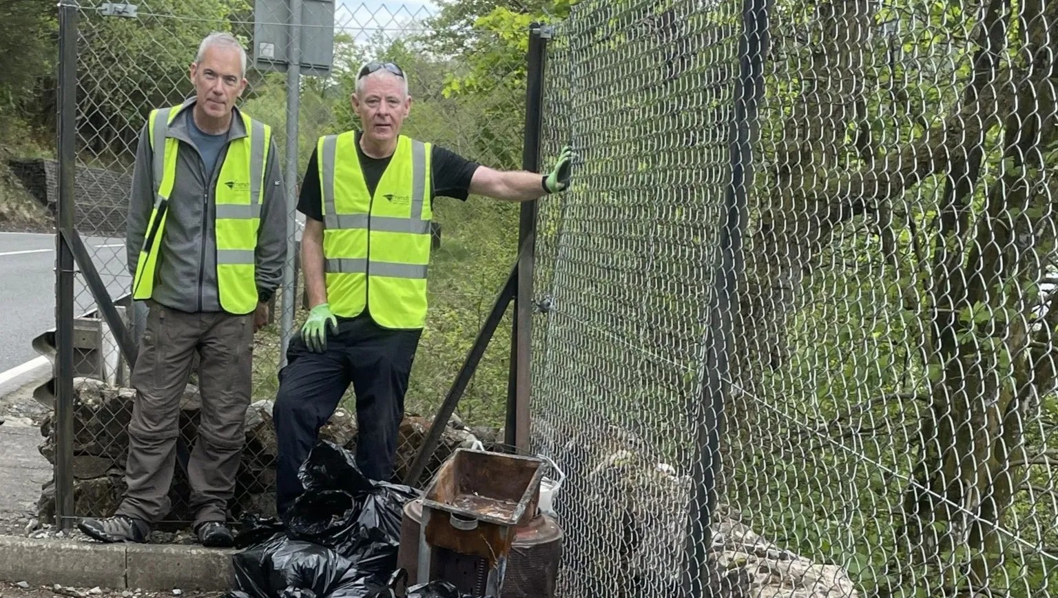 Two men stand behind a bunch of abandoned bags, a broken metal gate and some rusty old metal that have been abandoned in a layby