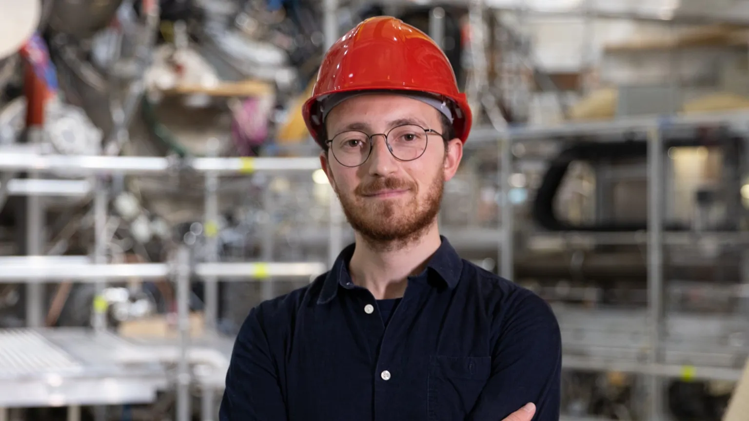 Proxima Fusion Wearing a red safety helmet and round glasses, Francesco Sciortino stands in front of fusion equipment.