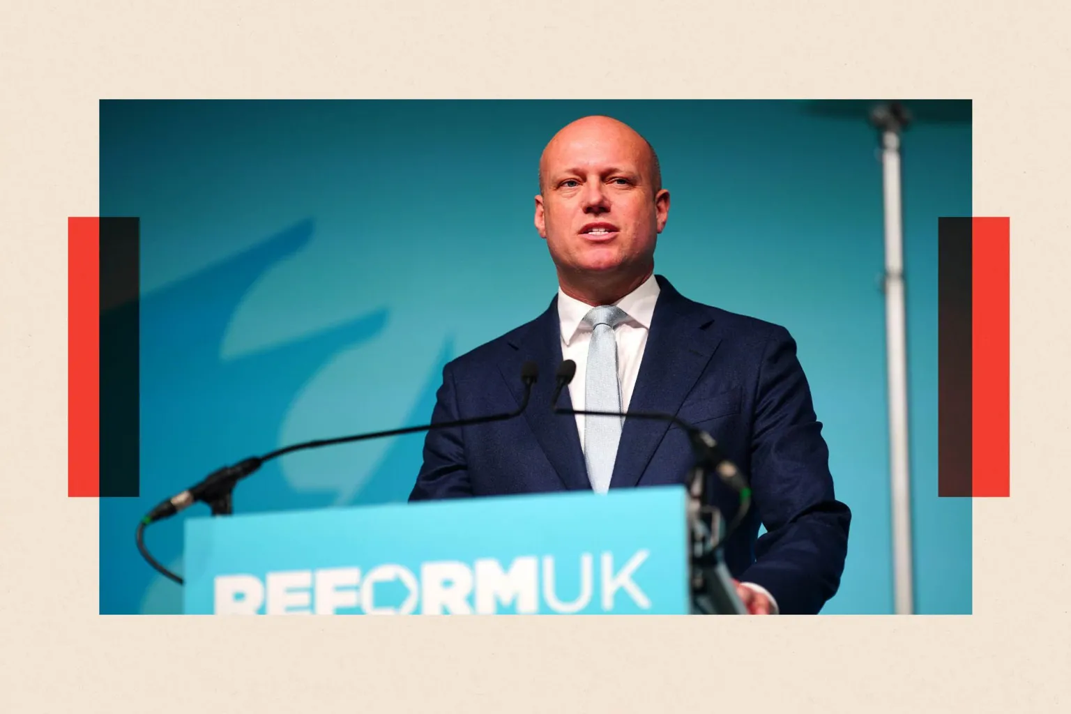  Reform UK leader in Wales Dan Thomas stands in front of a turquoise background on which the outline of a dragon is visible. In front of him is a lectern with a microphone and the Reform UK logo. He wears a dark shirt, a white shirt and a pale blue tie.